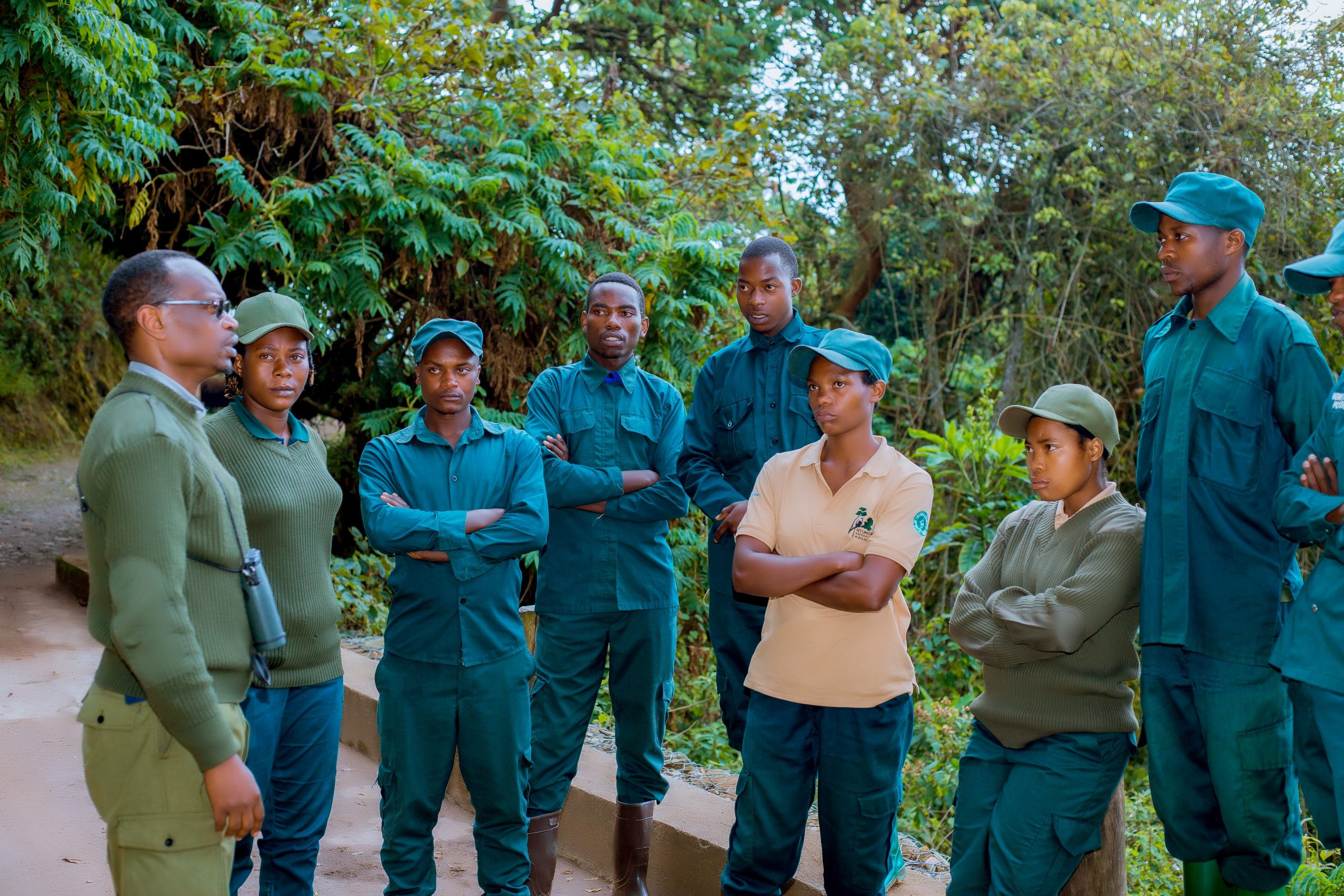 A group of park rangers and conservation trainees, dressed in green and beige uniforms, stand in a semi-circle outdoors in a forested area. Clever appears to be speaking or giving instructions, while the others listen attentively with arms crossed, surrounded by lush green foliage.
